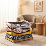 Pile de quatre lits pour chien et chat empilés sur un support en bois, dans un salon moderne avec un fauteuil bouclé et une décoration lumineuse. Les lits présentent différents motifs : rose à motifs abstraits, géométrique gris et rose, jaune à motifs de chiens, et bleu marine à motifs d'animaux.