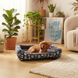 Chiot Cavalier King Charles Spaniel couché dans un panier pour chien douillet, bleu marine à motifs, dans un salon moderne avec des plantes et un fauteuil.
