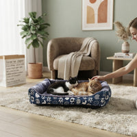 Panier douillet bleu à motifs pour chats dans un salon moderne et chaleureux, avec une femme caressant ses deux chats blottis dedans.
