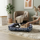 Panier douillet bleu à motifs pour chats dans un salon moderne et chaleureux, avec une femme caressant ses deux chats blottis dedans.