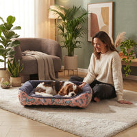 Panier pour chien rose à motifs dans un salon moderne et chaleureux, avec un Cavalier King Charles Spaniel endormi et une femme le caressant.
