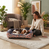 Panier pour chien rose à motifs dans un salon moderne et chaleureux, avec un Cavalier King Charles Spaniel endormi et une femme le caressant.