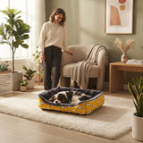 Panier pour chien jaune à motifs dans un salon chaleureux avec une femme debout à côté d'un chien endormi et des plantes vertes.