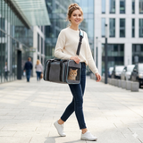 Une jeune femme en pull blanc et jean marche en ville avec un sac de transport pour animaux gris et bleu porté en bandoulière, laissant entrevoir un chat roux à travers le filet.