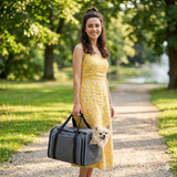 Une jeune femme en robe d'été jaune porte à la main un sac de transport gris pour animaux dans un parc, avec un petit chien blanc qui passe la tête par l'ouverture.